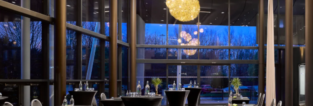 Inside a hotel meeting room with floor-to-ceiling-windows, with a meetings tables covered in black table clothes, and night time darkness outside the windows.