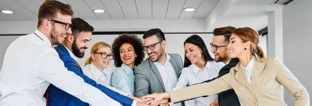 A group of people in formal business attire with their hands all together in the centre of the photo, all smiling to visually represent optimism for meetings.